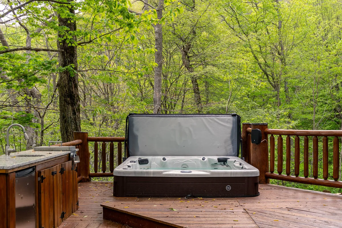 The wooden deck features a hot tub, surrounded by trees and nature, providing a spot for relaxation and leisure. The outdoor environment is both private and inviting, illustrating the kind of amenities guests and their pets can enjoy after a day on the trails.
