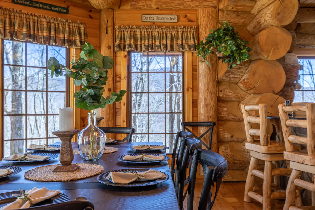 A dining area featuring a sturdy wood table set beneath exposed beams, with large windows letting in natural light and offering scenic views. The table comfortably seats a large group, surrounded by rustic charm and accented with a vase of greenery.