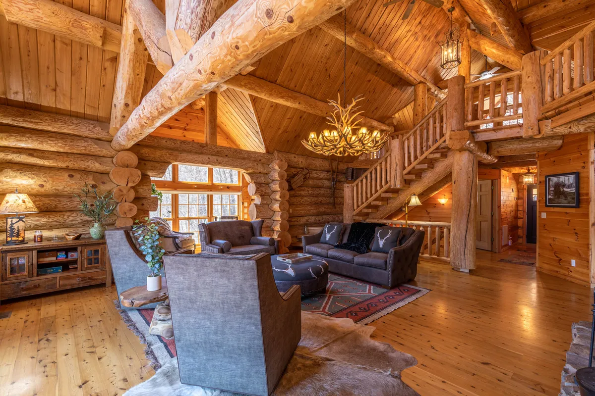 A spacious seating area with log walls, a chandelier, a rustic staircase, and large windows that overlook the natural surroundings. The space is designed for relaxation and social gatherings, with a warm, welcoming atmosphere enhanced by natural light and scenic views.