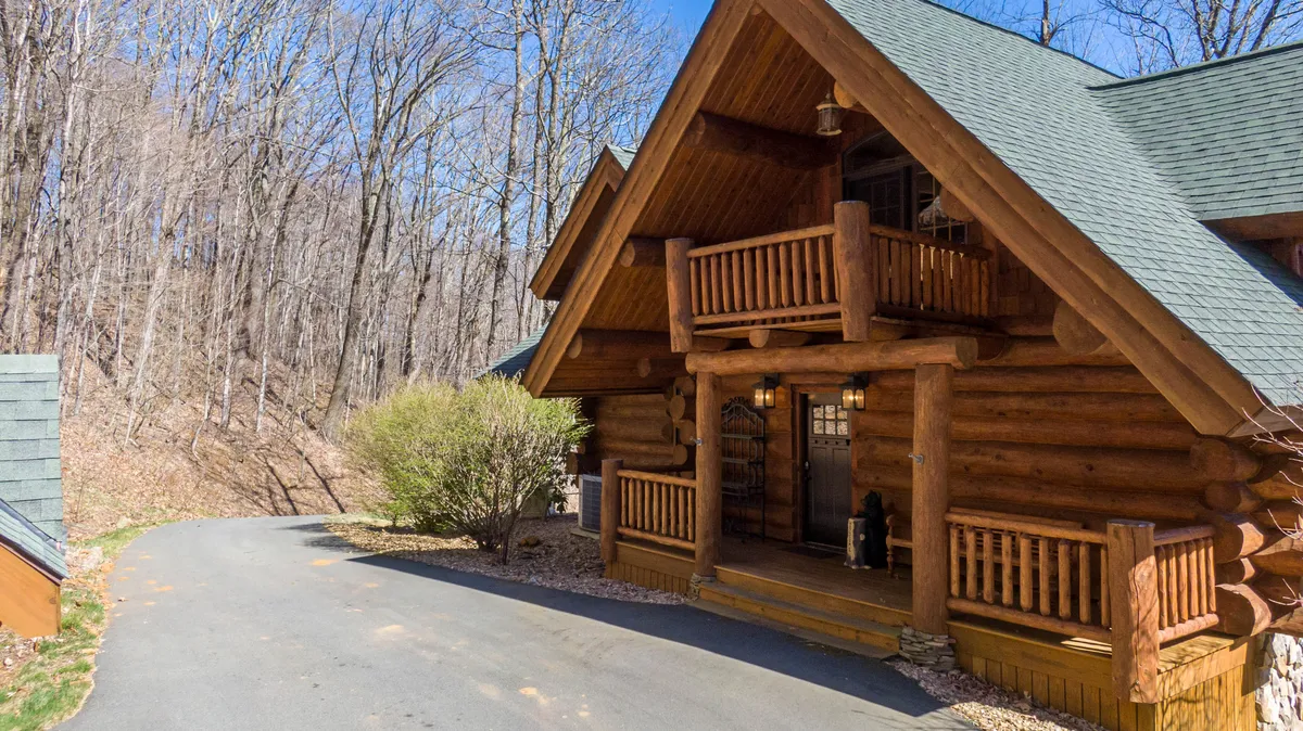 This photo presents the log cabin's exterior, complete with a wooden balcony and a driveway leading up to the lodge. The welcoming façade is nestled in its natural surroundings, offering a sense of arrival and outdoor adventure.