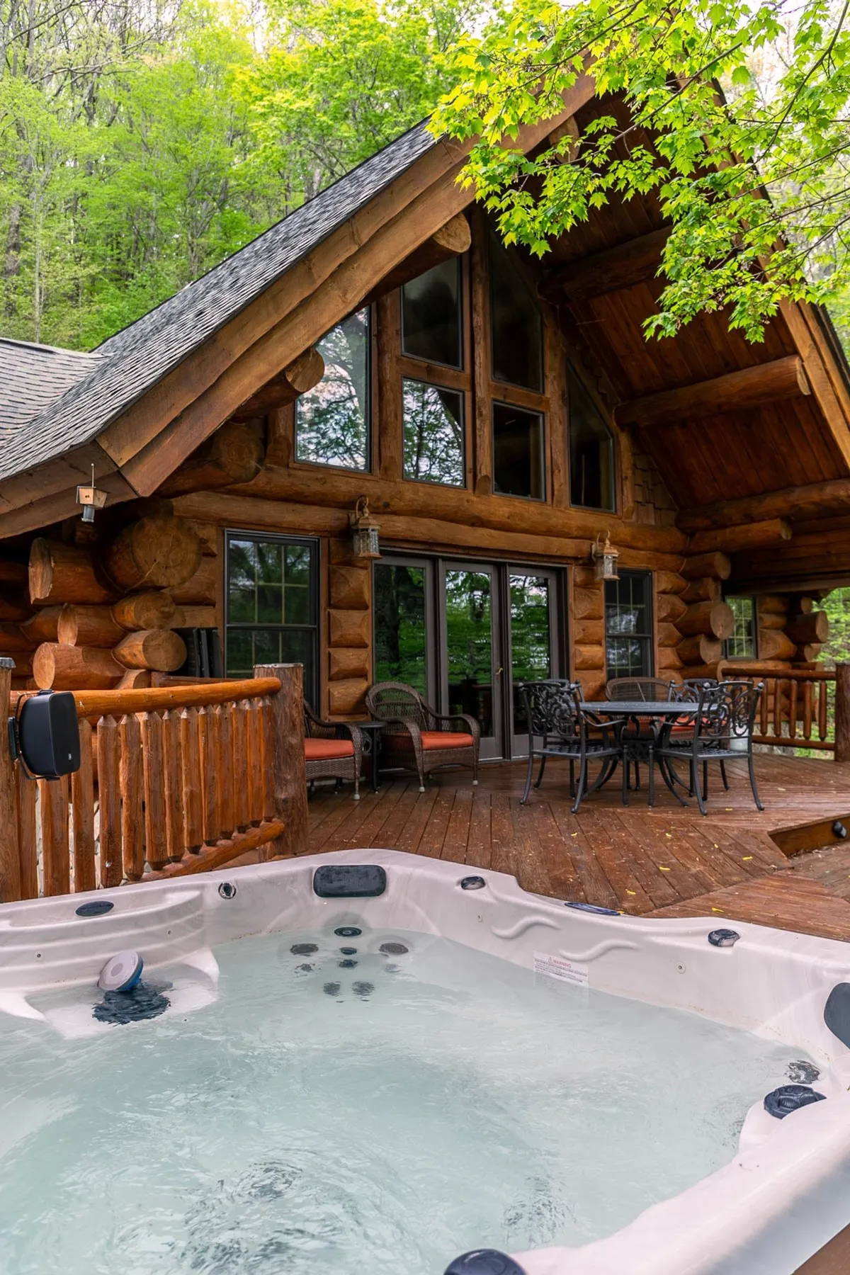 A wide exterior shot of the log cabin’s expansive wooden deck featuring a bubbling hot tub, cozy outdoor seating, and a dining area, all surrounded by lush trees. The log cabin structure and large windows create a seamless connection between indoor and outdoor living.