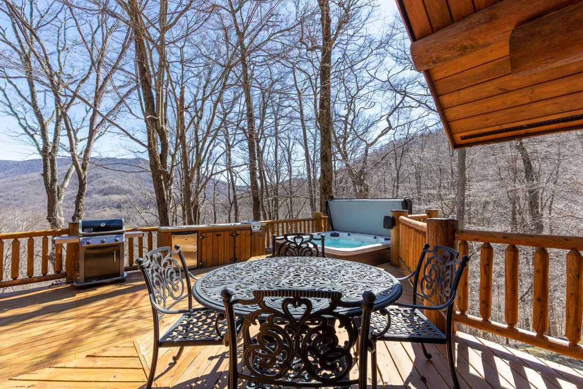 The image features an inviting outdoor living area with a spacious wooden deck, a hot tub, a large dining table, and a grill. The setting is surrounded by lush trees, creating a natural and secluded atmosphere, ideal for relaxation and group gatherings.