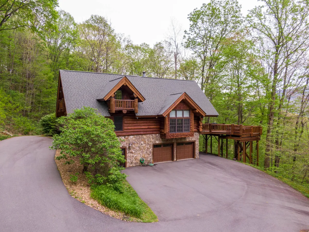 A rustic log cabin with a spacious deck, stone foundation, and lush trees surrounding the property. The setting communicates a direct connection to nature, inviting guests to relax, gather, or set out for nearby adventures in the mountains or on trails.