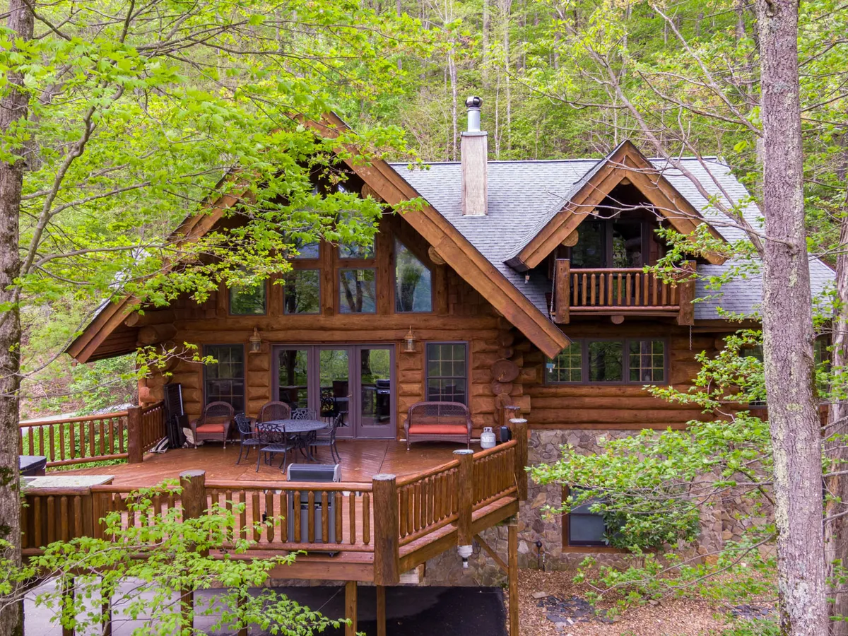 A log cabin with a generous deck surrounded by mature trees, inviting relaxation or small gatherings. The outdoor space feels integrated with the forest, encouraging exploration and quiet moments in nature.