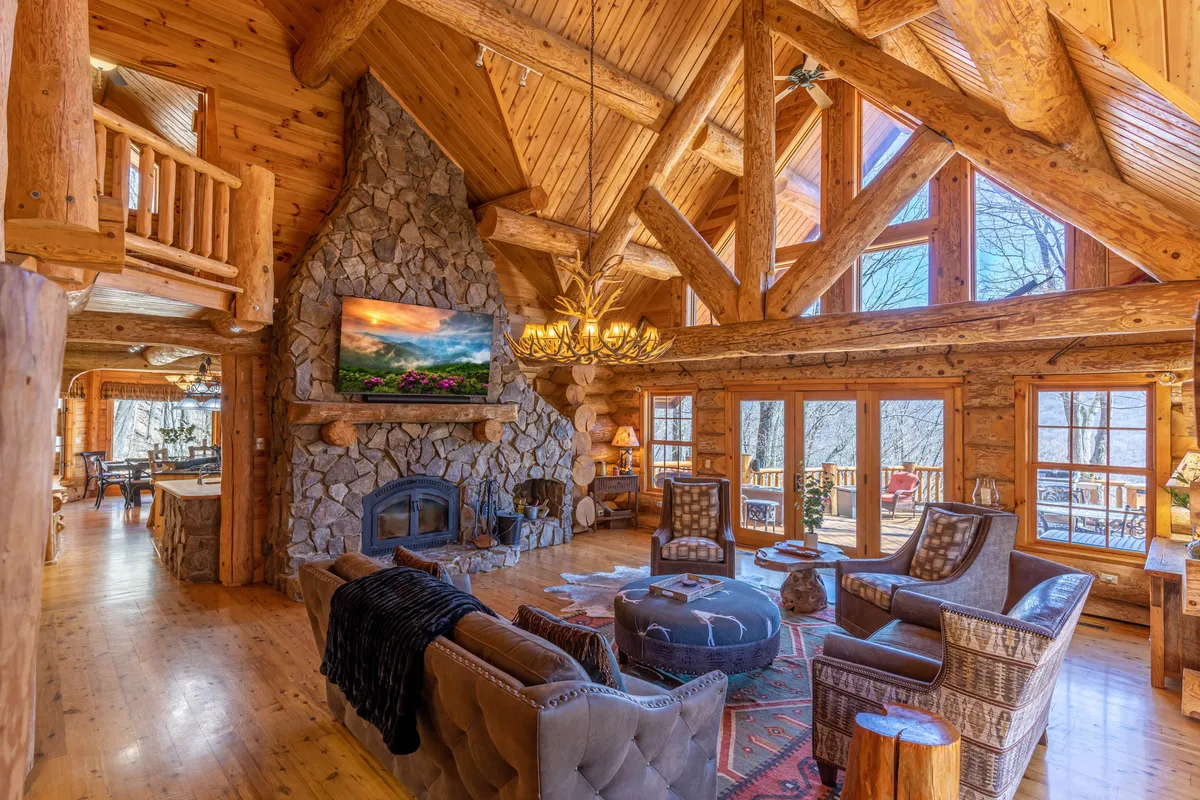 A spacious living room with a dramatic large stone fireplace stretching two stories high, surrounded by comfortable seating. Natural light floods in through floor-to-ceiling windows and wooden beams emphasize the vaulted ceiling. The cozy arrangement creates an inviting space for gatherings, relaxation, and entertainment.