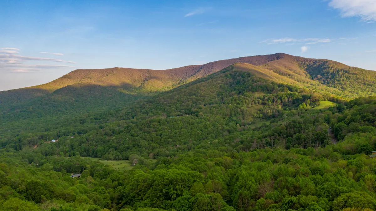 A sweeping view of rolling mountains, lush green trees, and a vast blue sky, offering a glimpse of the miles of private nature trails and the untouched wilderness that adjoins the property.
