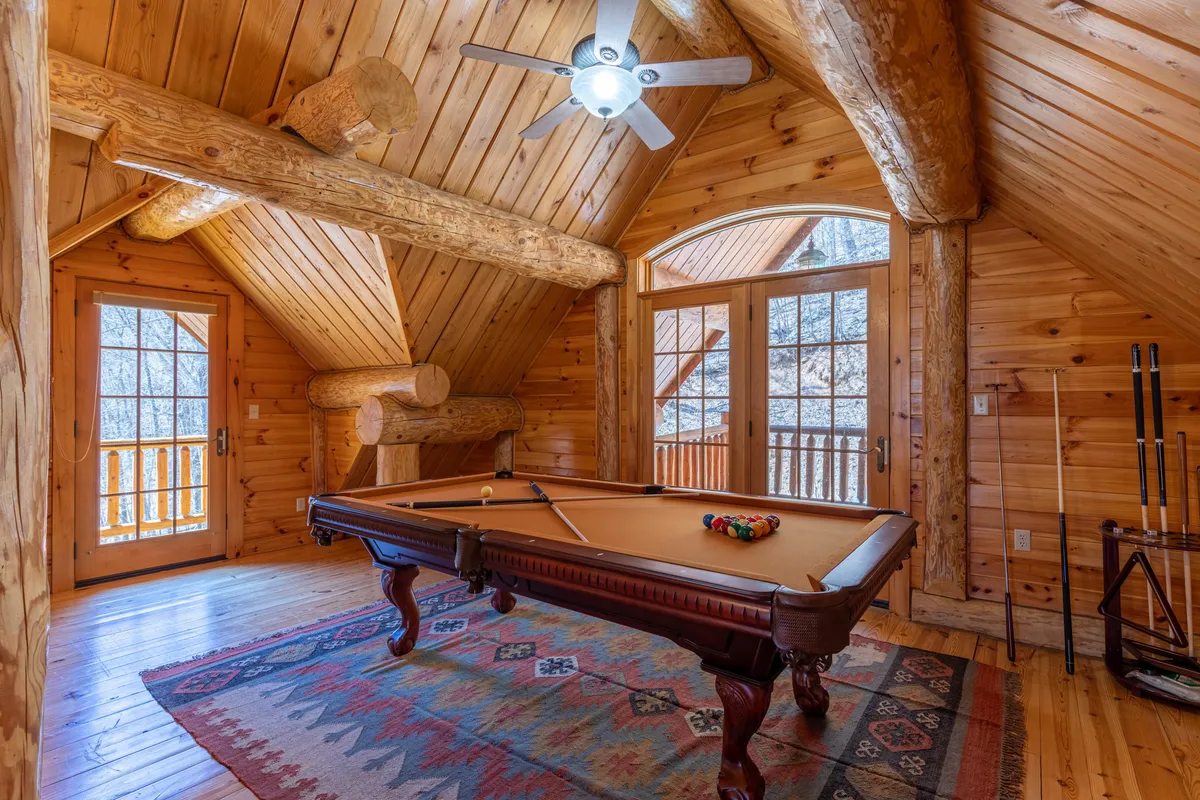 This image features an inviting entertainment loft with a pool table as the centerpiece, large windows flooding the space with natural light, exposed wooden beams, and a ceiling fan. The open design allows for both game play and relaxation, while the expansive windows offer glimpses of mountain views beyond.