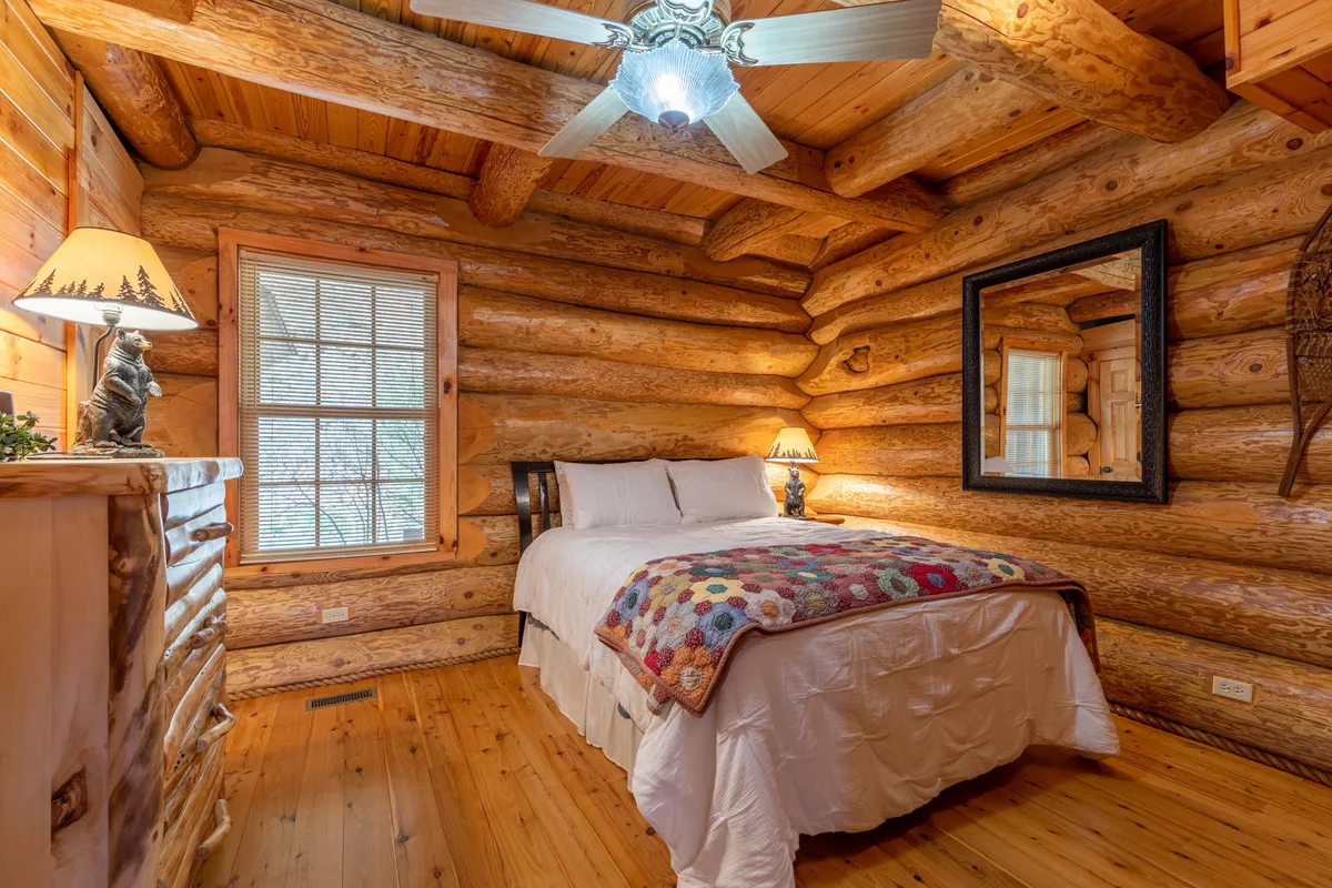 This image features a cozy bedroom with log walls, a comfortable bed adorned with a colorful blanket, a soft lamp casting a warm glow, a simple mirror, and rustic wooden furniture. Natural light filters in, enhancing the tranquil vibe of the space.