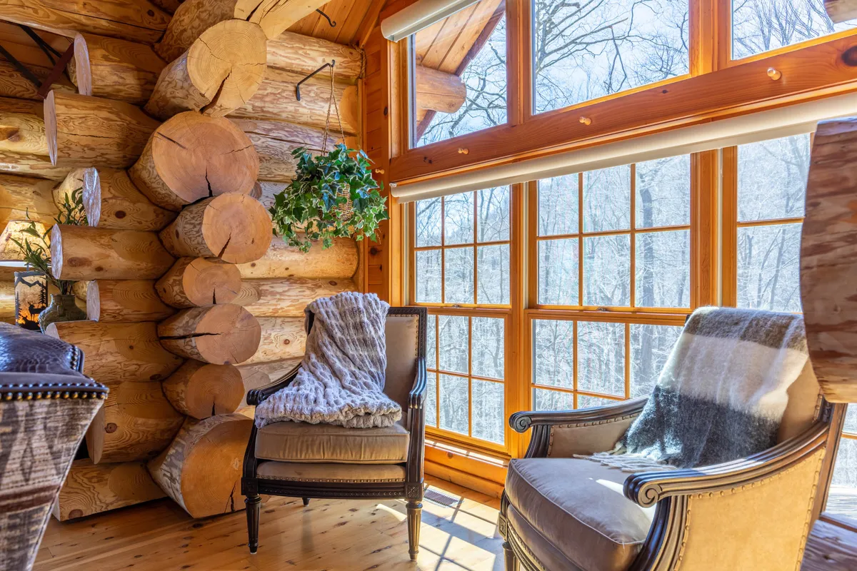 A cozy chair is positioned by large windows set into log cabin walls, with greenery visible outside. The sunlight pours through, creating an inviting nook ideal for reading or relaxing while connected to the outdoors. The overall feel is tranquil, blending the comfort of indoor space with the inspiration of mountain views.