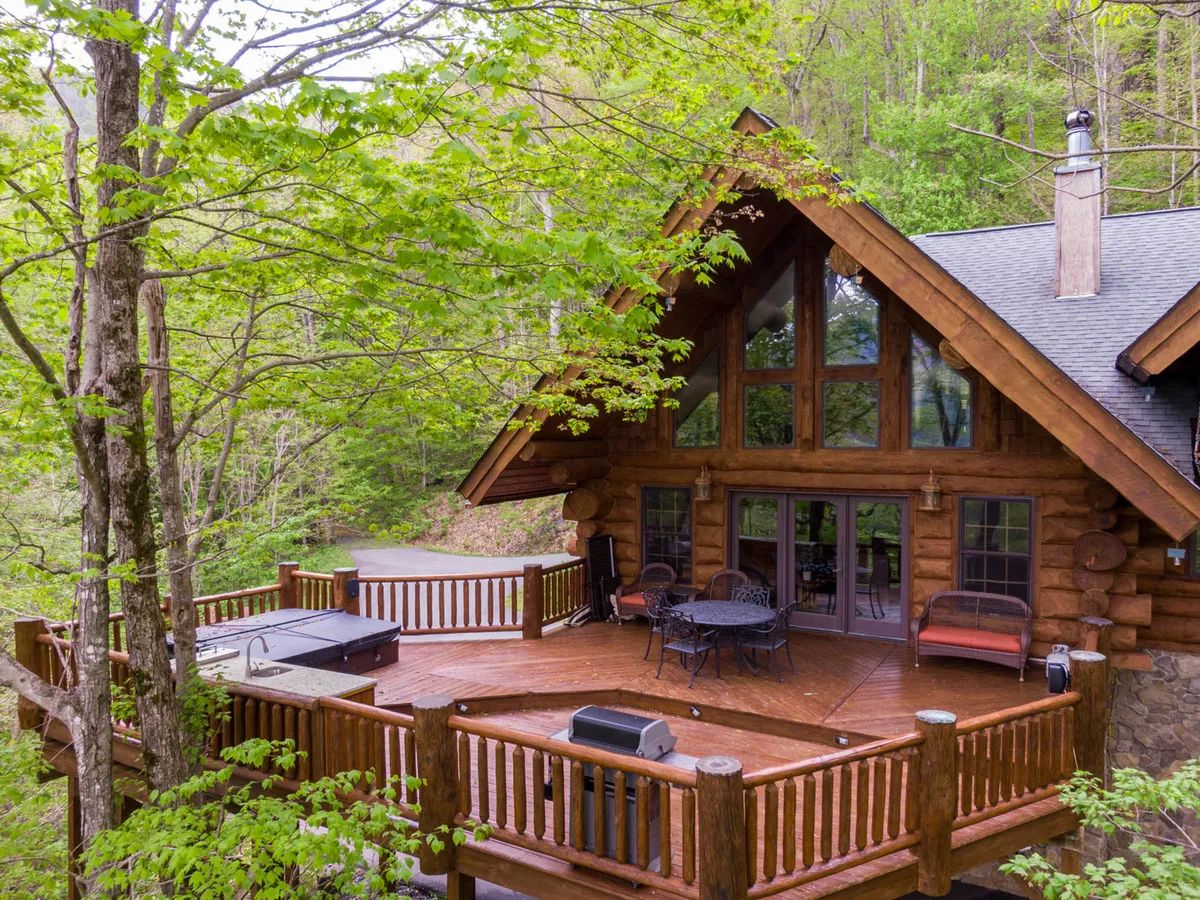 This image presents the log cabin's exterior with a spacious deck surrounded by lush forest. Large windows offer views both in and out, and the deck area suggests room for relaxation or gatherings. The scene evokes a seamless transition from the comfort of the lodge to the natural world just steps away, with hints of the trails and wildflower-filled forest beyond.