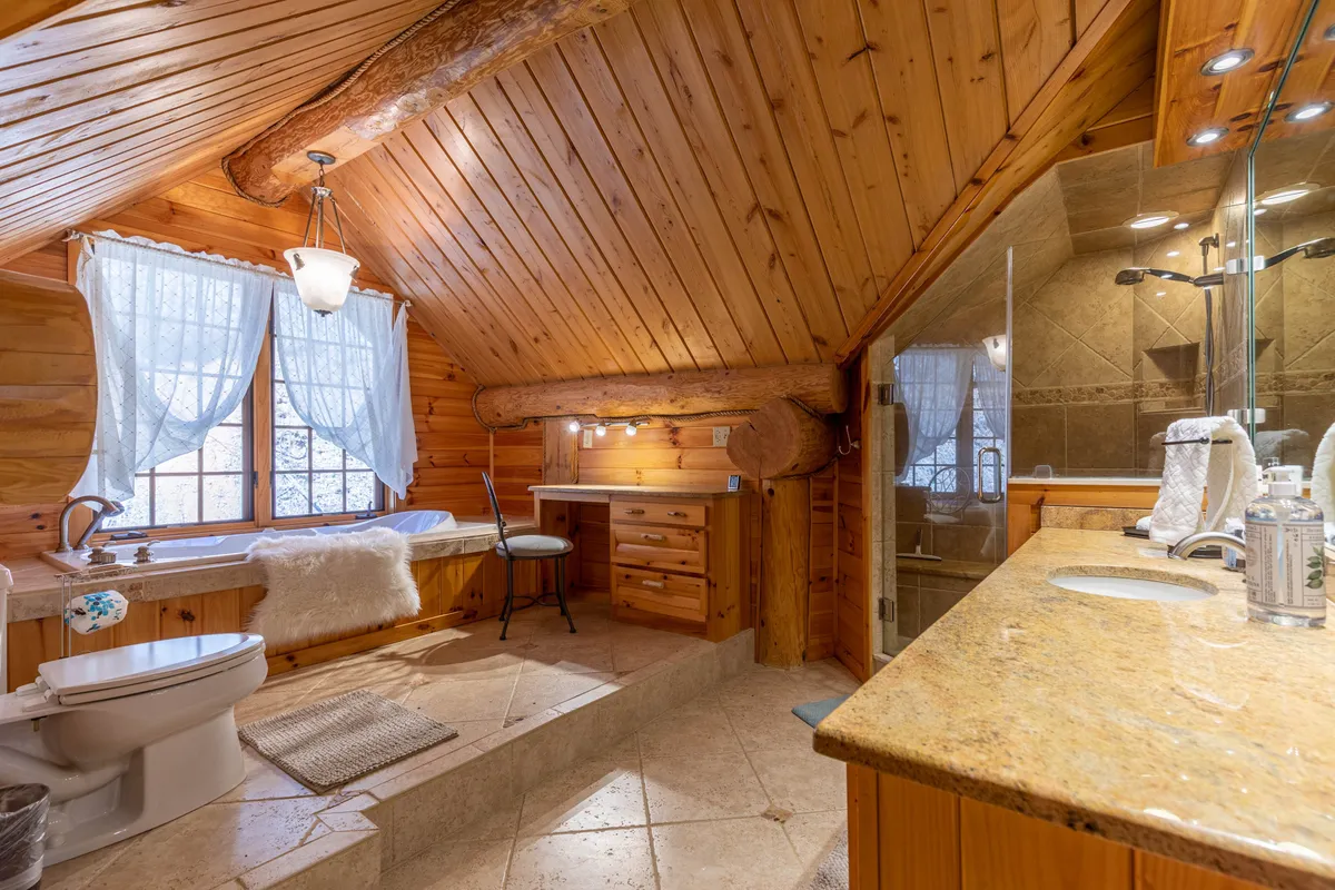 A luxurious bathroom scene highlighting a deep soaking tub with a view, wooden beams, large windows letting in natural light, and a natural stone countertop at the vanity. The tranquil atmosphere suggests opportunities for relaxation and solitude.