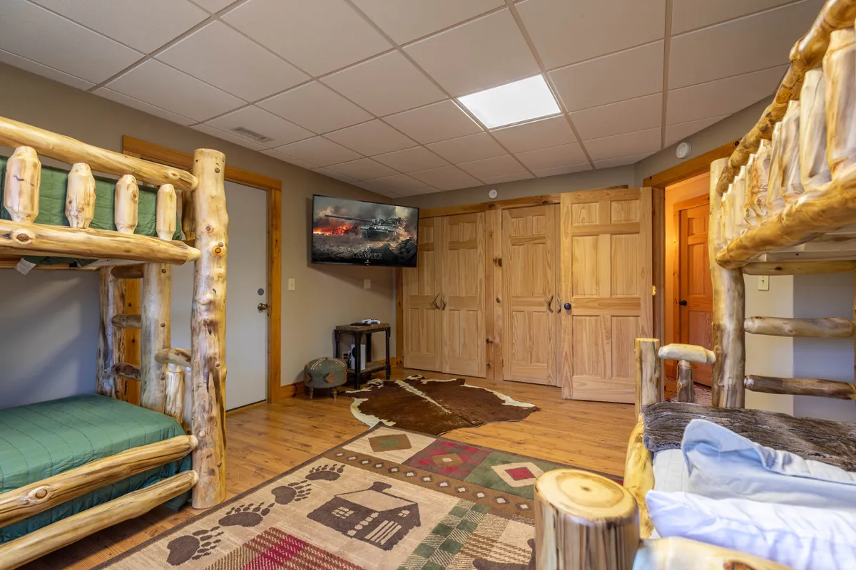 A fun and inviting bunk room, complete with log bunk beds, a television with a scenic backdrop, wooden storage cabinets, and a playful rug with animal prints. The space feels communal and welcoming, ideal for kids or groups.