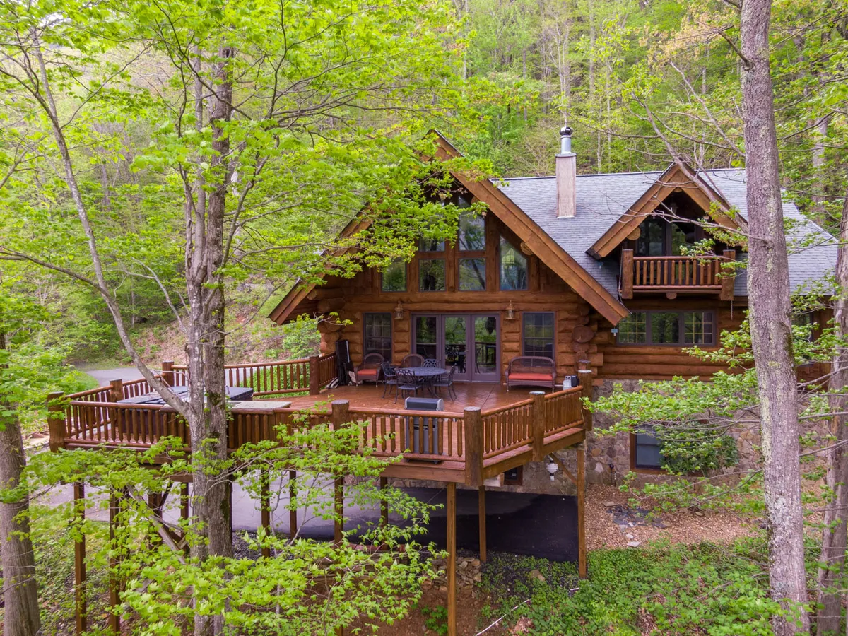 A peaceful log cabin featuring a large wooden deck overlooking vibrant greenery and mature trees. The scene radiates tranquility, with expansive windows and outdoor space ideal for morning coffee or gathering after exploring local attractions.