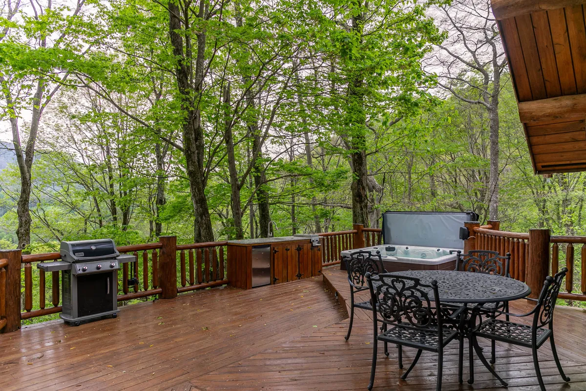 A bright view of an outdoor dining table and grill station next to a bubbling hot tub on the deck, with tall trees providing privacy and a natural backdrop. The space feels ready for al fresco meals and gatherings.