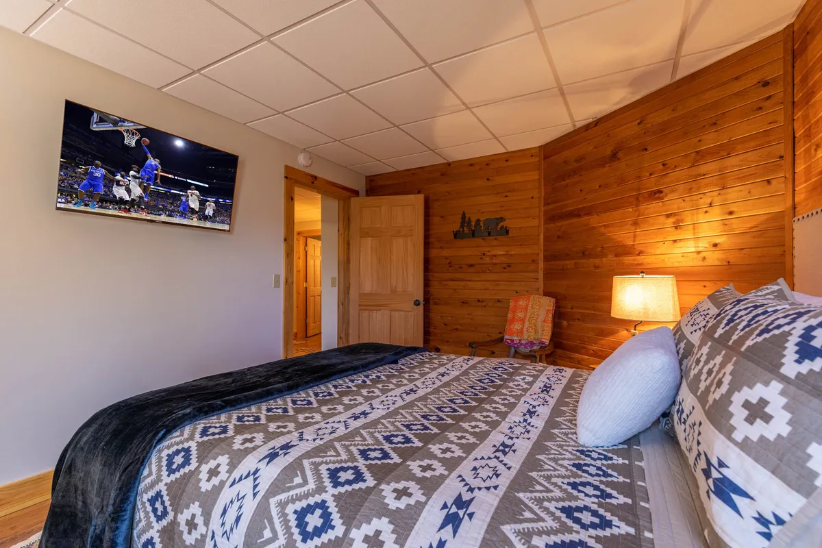 A well-appointed guest bedroom featuring a bed with patterned bedding, a wall-mounted television, warm wood paneling, a softly-glowing lamp, and a decorative chair. The room exudes comfort and modern amenities.