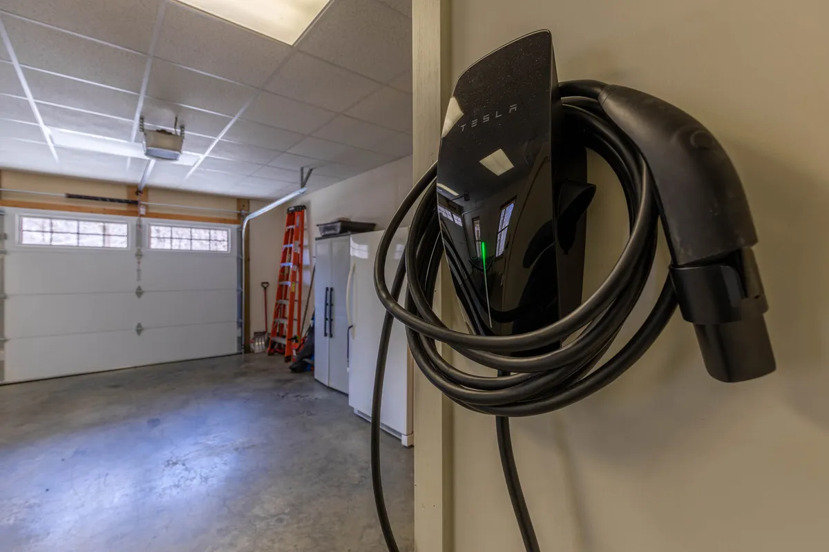 A Tesla charger mounted on the wall inside a tidy, well-lit garage with tools and storage equipment visible. The space demonstrates modern amenities and support for electric vehicles, emphasizing convenience and sustainability for guests.